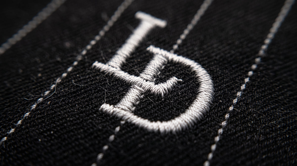 Détail macro du monogramme LD brodé en fil argenté sur le pantalon noir rayé